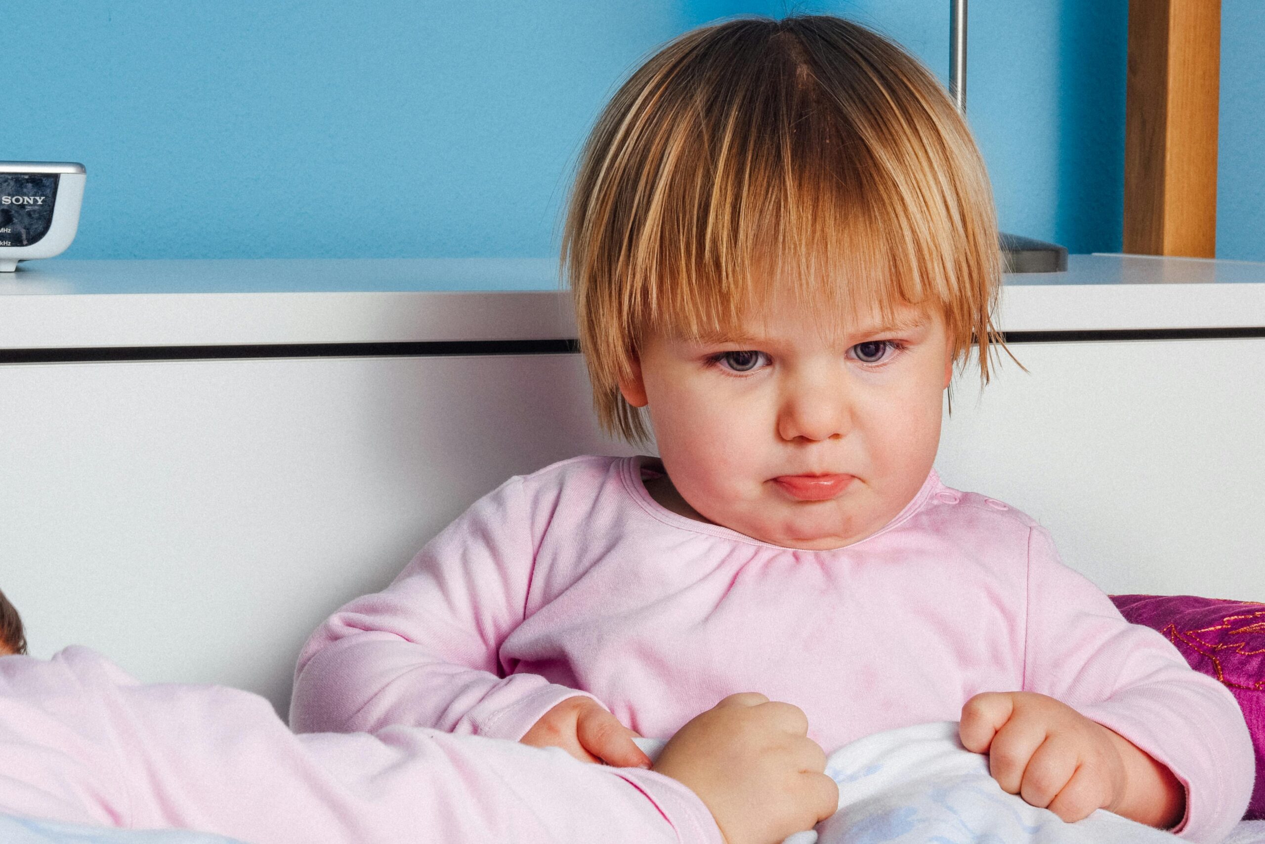 Cute toddler with a pout in a pink outfit indoors, showcasing an expressive mood.